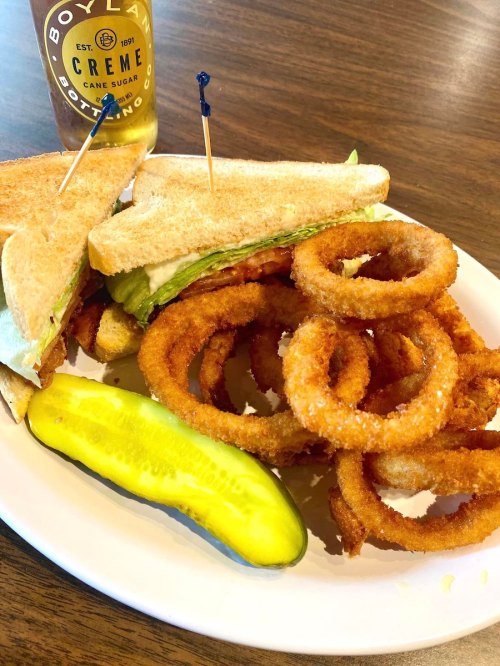 A plate with a sandwich, onion rings, and a pickle, with a drink in the background.