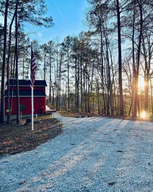 A gravel driveway leads to a red house surrounded by tall trees, with a flagpole and a sunset reflecting on a nearby lake.