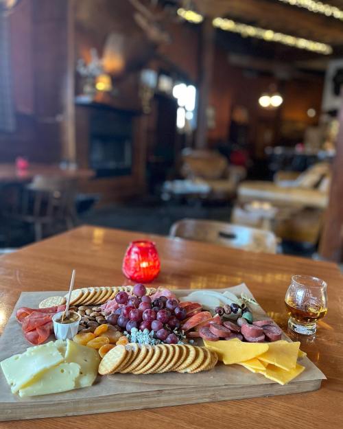 A wooden platter with assorted cheeses, meats, grapes, crackers, and a glass of whiskey, set in a cozy, rustic lounge.