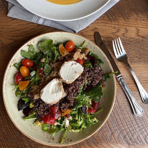 A plate of salad with mixed greens, cherry tomatoes, quinoa, and sliced chicken, with a bowl of soup in the background.