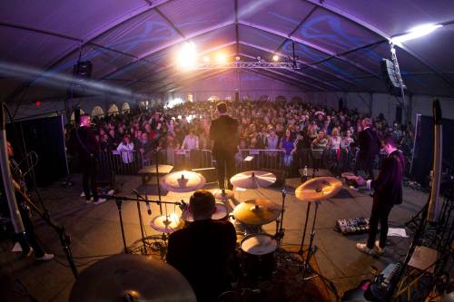 A lively concert scene with a large audience under a tent, viewed from the stage with musicians in the foreground.