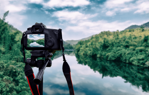 A camera on a tripod captures a serene river scene surrounded by lush greenery and mountains under a cloudy sky.