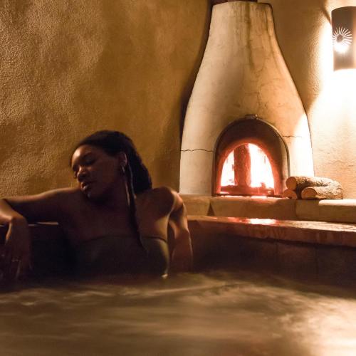 A woman relaxes in a hot tub beside a glowing fireplace, creating a cozy and serene atmosphere.