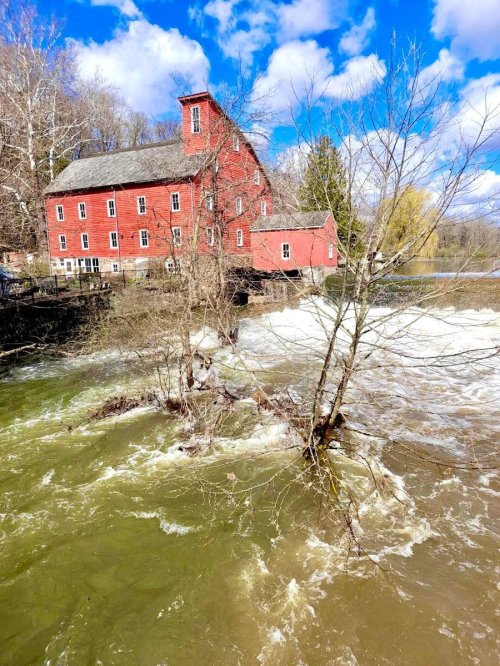 A red historic building beside a rushing river, with trees and blue skies in the background.