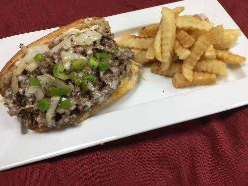 A plate featuring a loaded sandwich with beef, onions, and green peppers, accompanied by crinkle-cut fries.