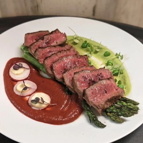 A plate of sliced steak served with asparagus, creamy sauce, and vibrant green puree, garnished with microgreens.