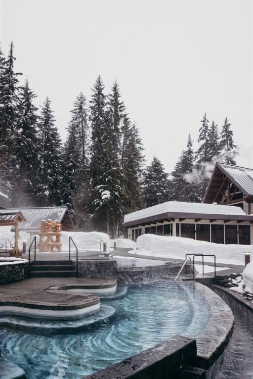 A serene hot spring surrounded by snow-covered trees and a cozy lodge in a winter landscape.
