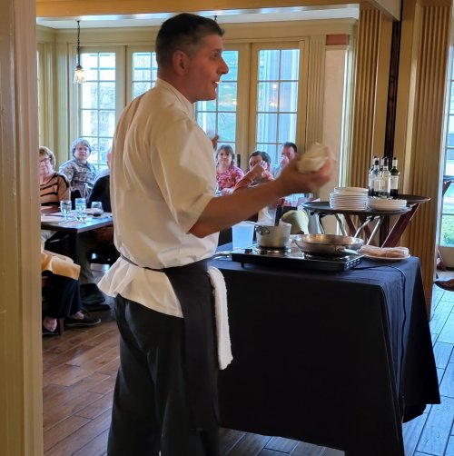 A chef demonstrates cooking techniques in a restaurant, with an audience watching attentively in the background.