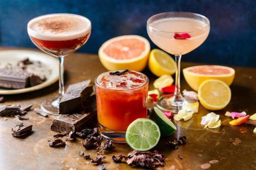 Three colorful cocktails on a table with chocolate, citrus fruits, and flower petals scattered around.