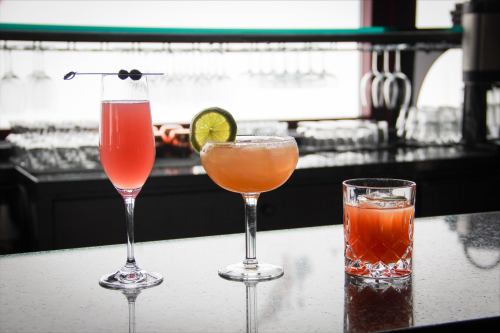 Three colorful cocktails on a bar counter: a pink drink in a flute, a margarita with lime, and a whiskey drink in a glass.