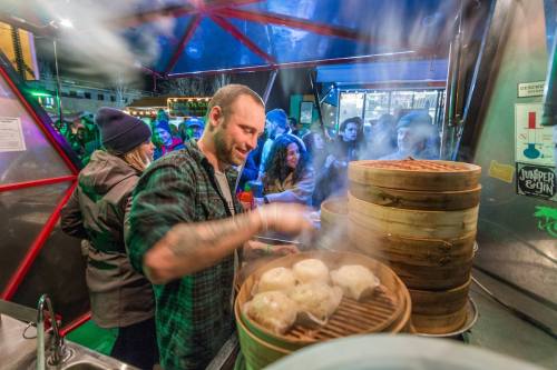 A chef prepares steaming dumplings at a busy food stall, with a crowd enjoying the atmosphere in the background.