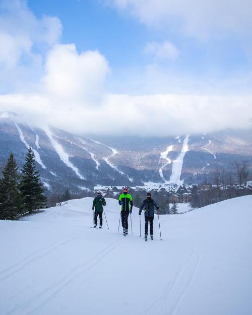 Three skiers traverse a snowy landscape with mountains and a village in the background under a partly cloudy sky.