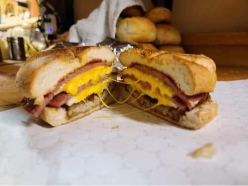 A close-up of a sliced sandwich with layers of cheese, ham, and sausage on a toasted bun, resting on a countertop.
