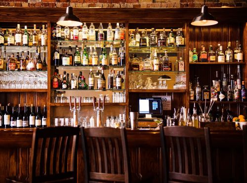 A well-stocked bar with various bottles of liquor, glasses, and a computer, set against a rustic wooden backdrop.