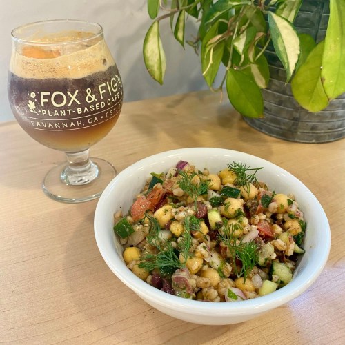 A bowl of colorful grain salad with vegetables and herbs, next to a glass of dark beverage, on a wooden table.