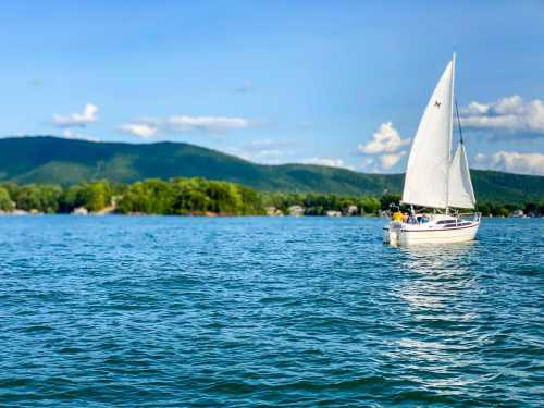 A sailboat glides across a calm lake, surrounded by green hills and a clear blue sky with fluffy clouds.