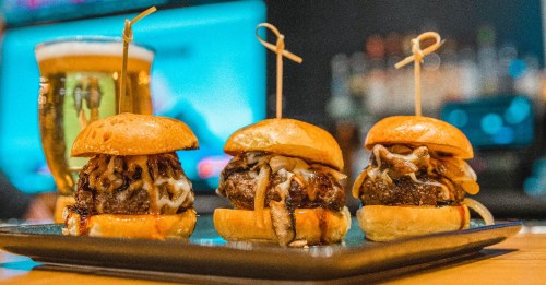 Three mini burgers with melted cheese and toppings on a black plate, accompanied by a glass of beer in the background.