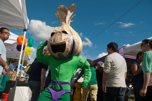 A person in a large onion costume poses playfully at an outdoor festival, surrounded by colorful tents and attendees.