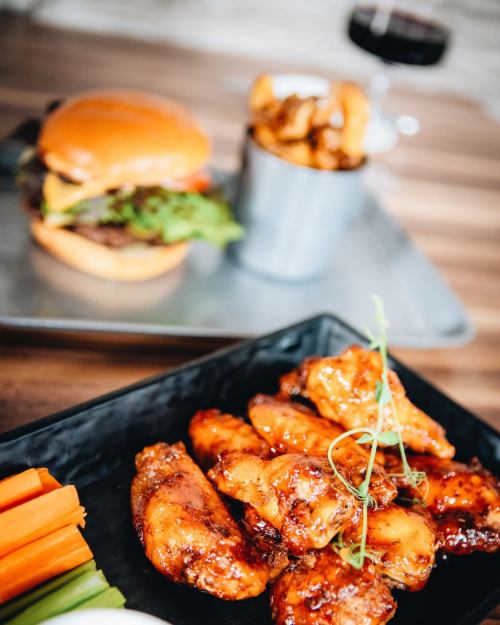 A plate of glazed chicken wings with celery sticks, alongside a burger and a cup of fries in the background.