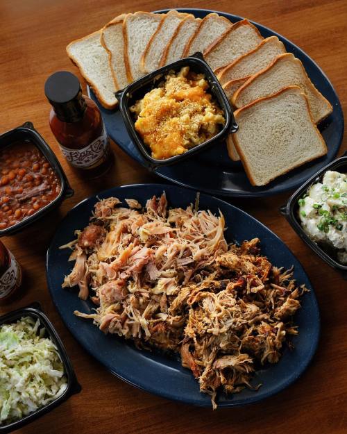 A platter of pulled pork, sides of coleslaw, baked beans, macaroni and cheese, and slices of bread with barbecue sauce.