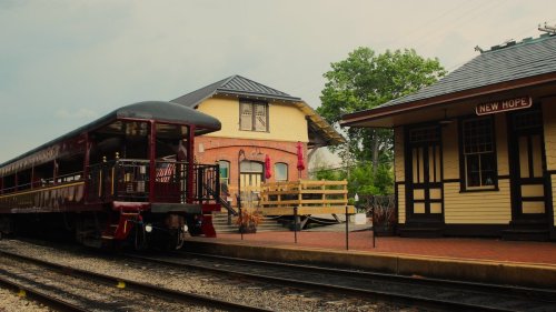 A vintage train at a quaint station with two distinct buildings, surrounded by greenery and a cloudy sky.