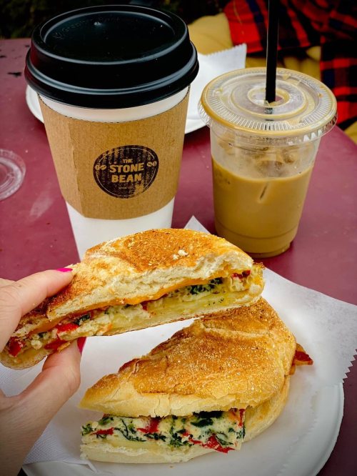 A hand holding a toasted sandwich with spinach and red peppers, alongside a coffee cup and iced coffee.