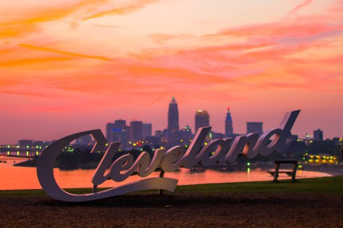 A large "Cleveland" sign in the foreground with a colorful sunset and city skyline in the background.
