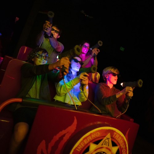 A group of four people in 3D glasses, holding toy guns, enjoying a ride in a dark, colorful arcade setting.