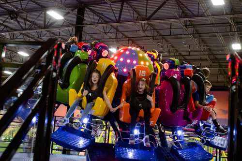 Two excited children on a colorful amusement ride, holding hands and smiling, with other riders in the background.