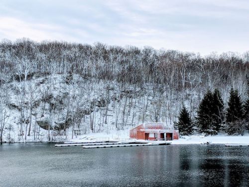 A snowy landscape featuring a red building by a calm lake, surrounded by trees and a snow-covered hillside.