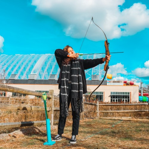 A person aiming an arrow with a bow outdoors, wearing a black outfit and standing near a colorful building.