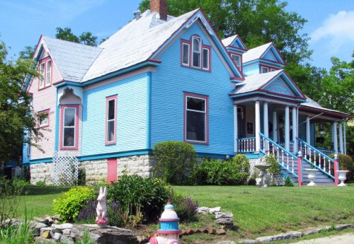A colorful Victorian-style house with blue siding, white trim, and a lush green yard featuring decorative elements.