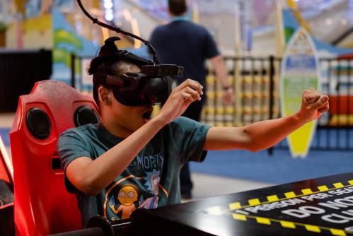 A child wearing a VR headset sits in a ride, excitedly reaching out with arms in a playful environment.
