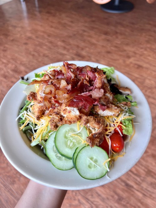 A bowl of salad topped with crispy chicken, bacon, cheese, and fresh vegetables like cucumbers and cherry tomatoes.
