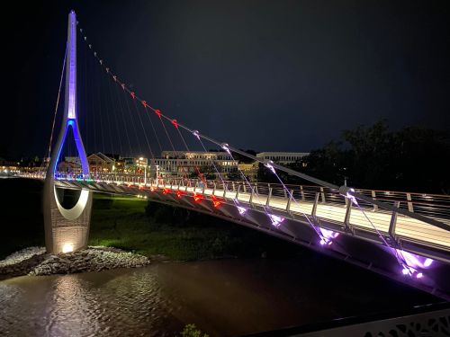 A brightly lit suspension bridge at night, featuring colorful lights and a serene river below.
