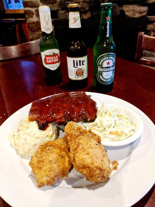 A plate of fried chicken, coleslaw, potato salad, and barbecue ribs, with three bottles of beer in the background.