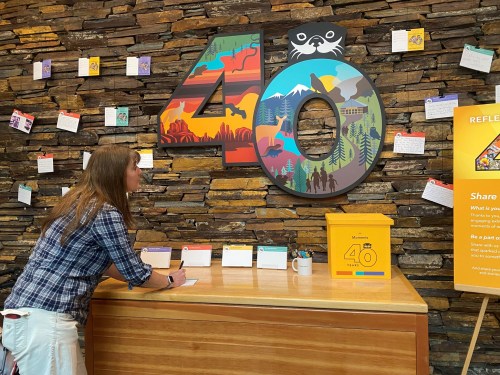 A person writes at a wooden table in front of a colorful "40" display against a stone wall, with notes and a yellow box nearby.