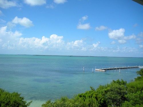 A serene coastal view featuring calm turquoise waters, a wooden pier, and a clear blue sky with scattered clouds.