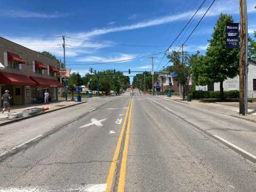 A wide, empty street lined with trees and buildings, under a clear blue sky with a few pedestrians in the distance.