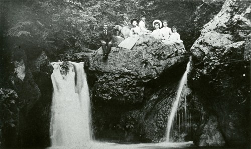 A vintage photo of a group of people sitting on rocks by a waterfall, surrounded by lush greenery.