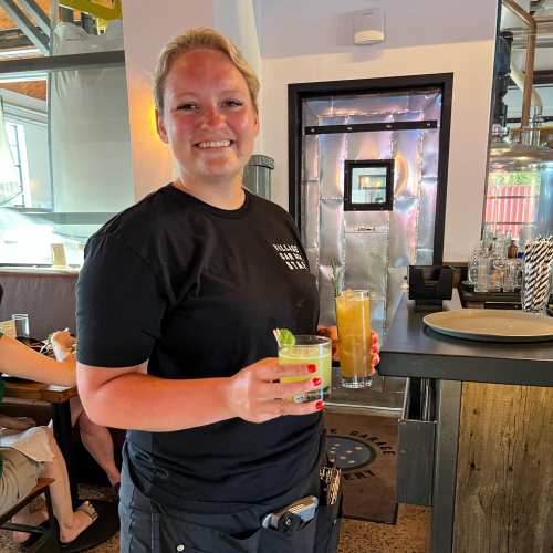 A smiling server holds two colorful cocktails in a restaurant setting.