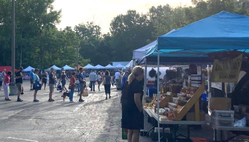A bustling outdoor market with vendors under blue tents and shoppers browsing various goods in a sunny setting.