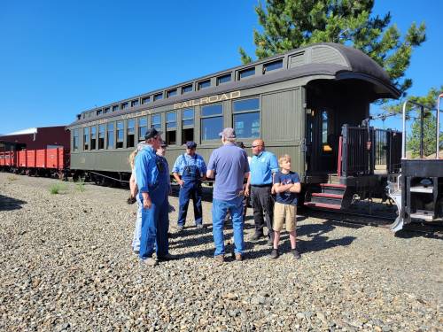 A group of people gathers near a vintage railroad car on a sunny day, surrounded by gravel and trees.