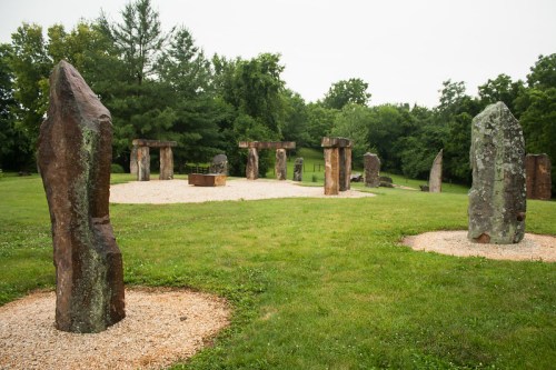 A grassy area featuring large standing stones arranged in a circle, surrounded by trees and a gravel path.