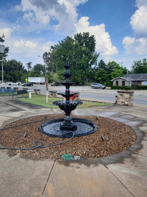 A black fountain surrounded by gravel, with trees and a cloudy sky in the background.