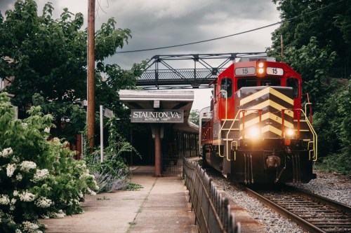 A red train approaches a station sign reading "Staunton VA" under a cloudy sky, surrounded by greenery.