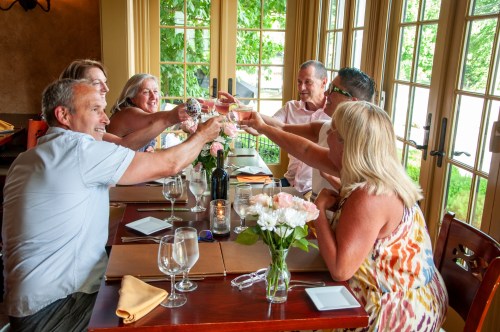 A group of six people toasting with drinks at a dining table, surrounded by flowers and a warm, inviting atmosphere.