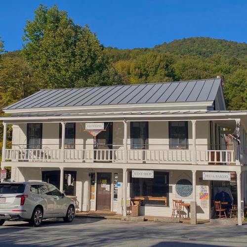 A charming two-story building with a metal roof, featuring a clothing store, wine shop, and deli/bakery, surrounded by trees.