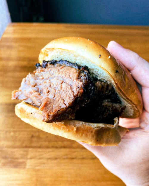A hand holding a juicy brisket sandwich with a soft bun, set against a wooden table background.