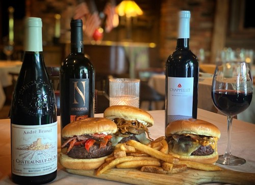 Two gourmet burgers with fries, flanked by three bottles of red wine and a glass of wine on a table.